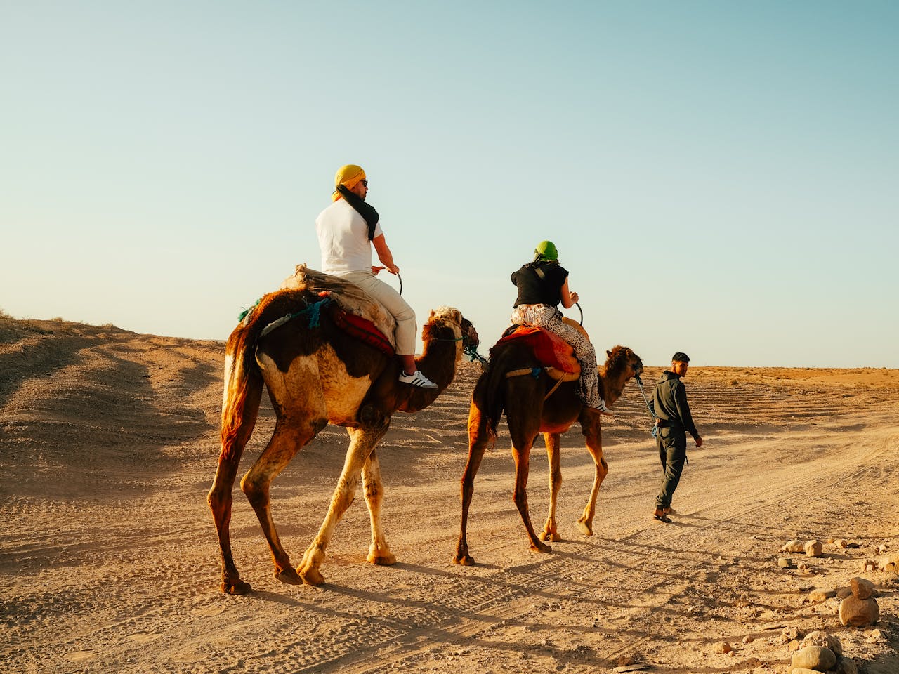 services-03 Two tourists riding camels in the vast Moroccan desert under clear blue skies.